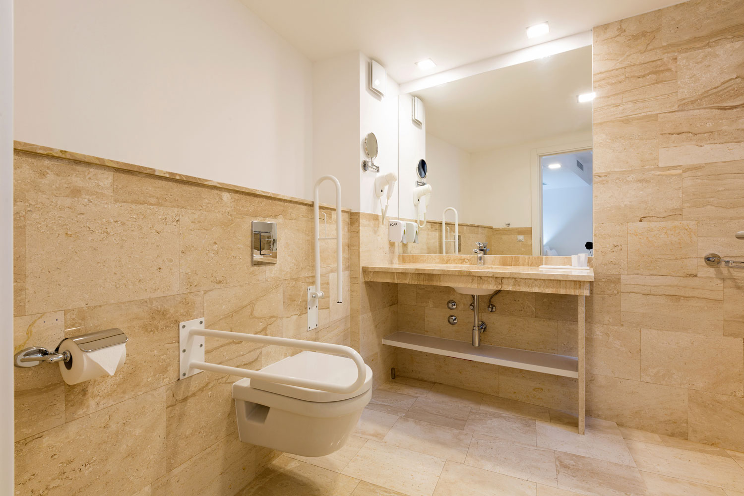 Salle de bains PMR intégrale en travertin beige avec barres d'appui blanches et vasque suspendue à Chilly-Mazarin.